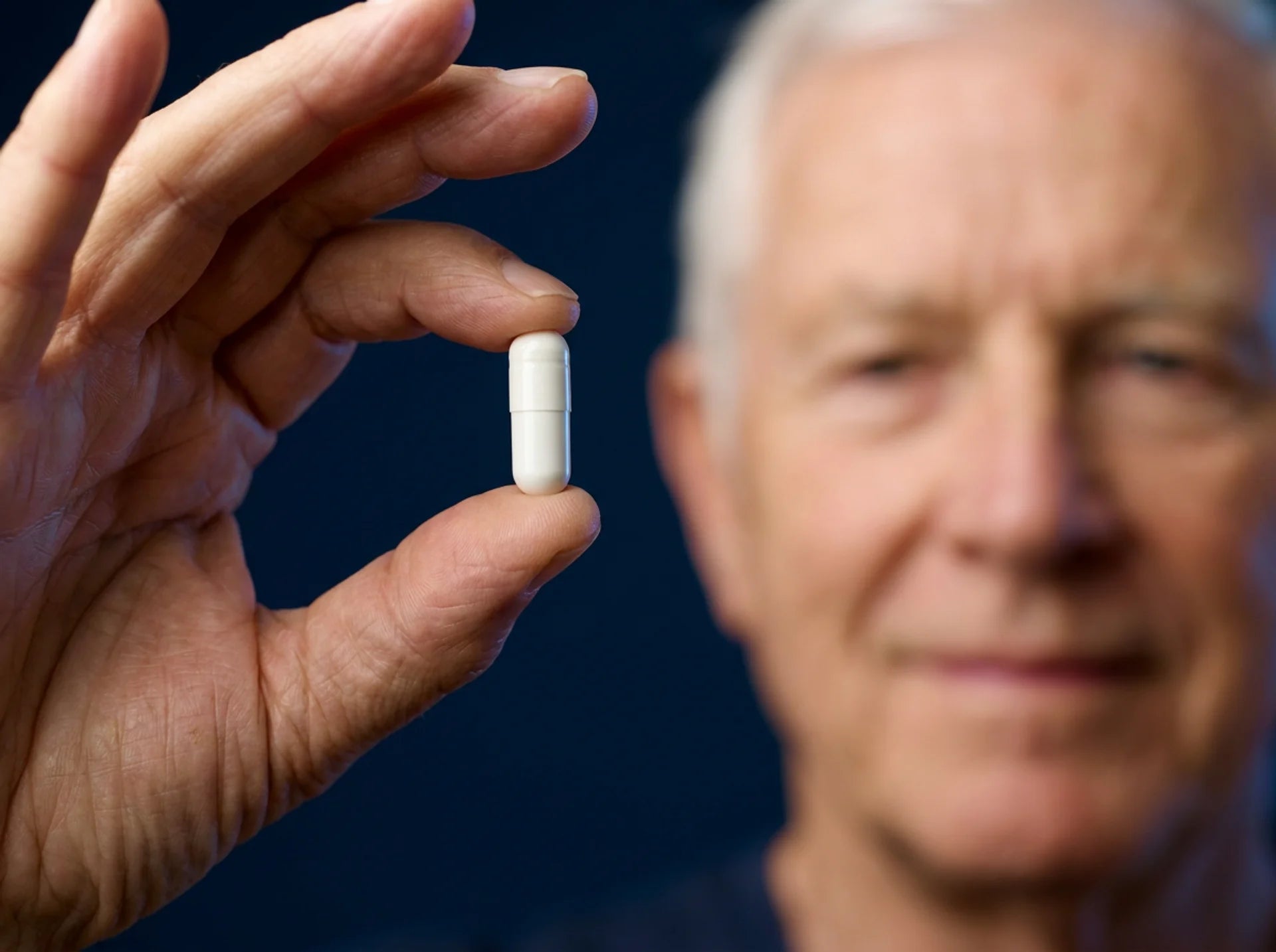 Illustrative image: an older man’s hand holding one white capsule, with a blurred face behind it